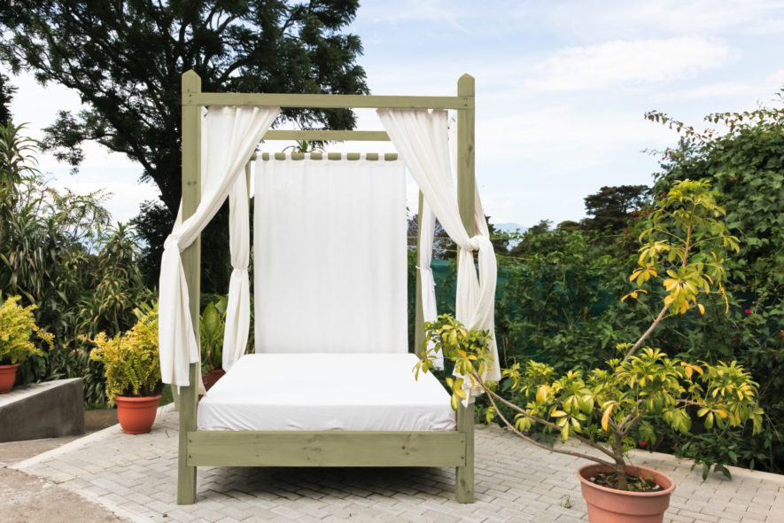 A wooden outdoor bed with white linens and privacy curtains on a brick patio at a tropical resort.